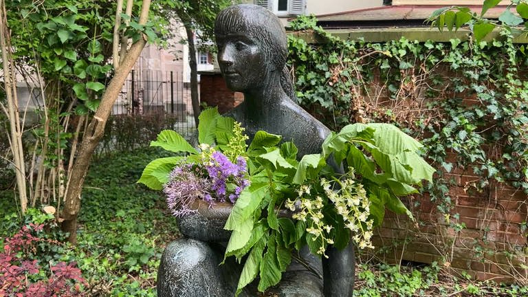 Man sieht eine Skulptur in Tübingen. Man nennt sie: die "Sitzende". Sie hat Blumen in den Arm gelegt bekommen. Viele davon sind grün. Man erkennt aber auch einen kleinen lila Strauß. (Foto: SWR)